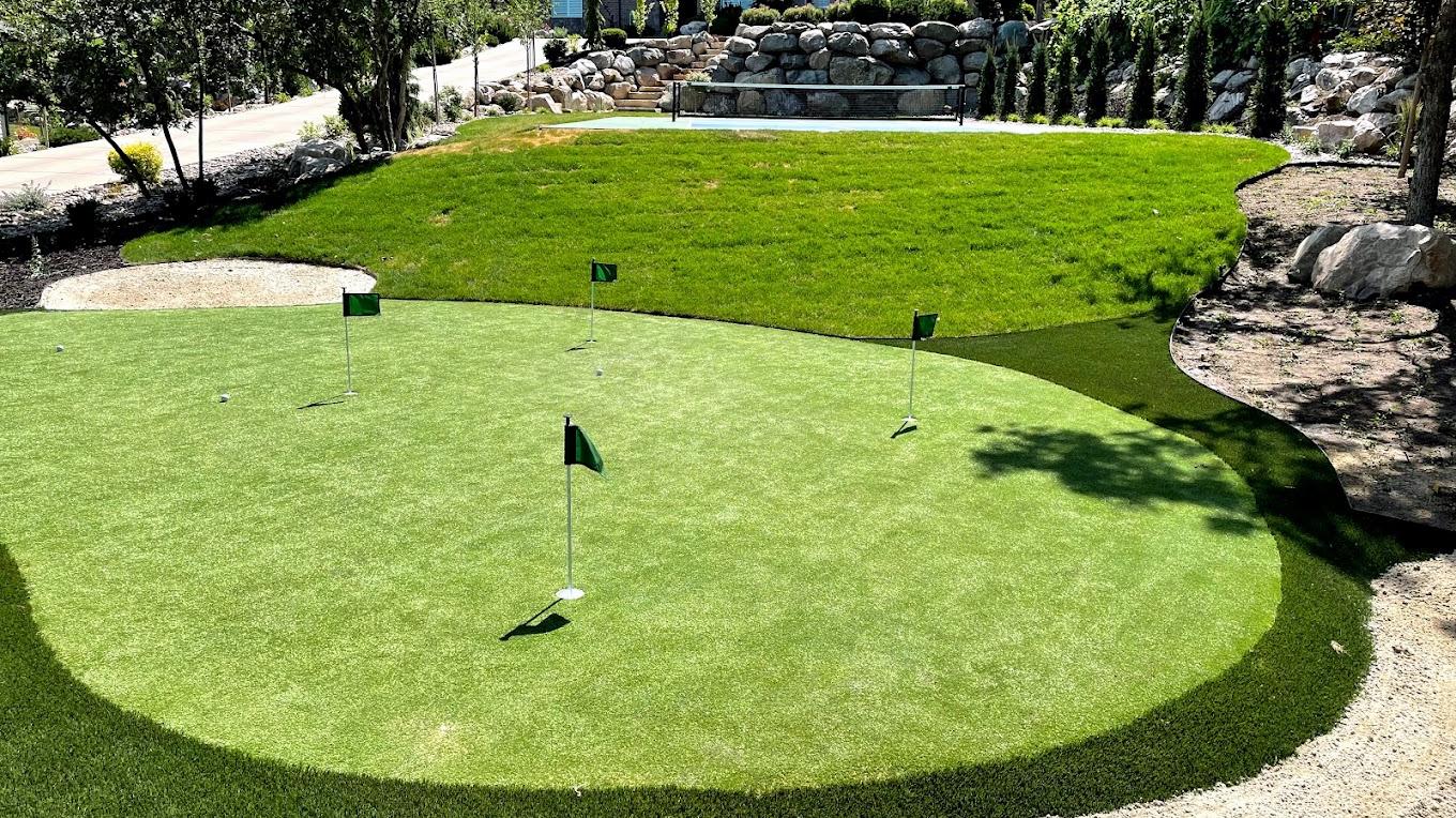 Backyard putting green with five flagsticks beside lawn, trees, and rock landscaping