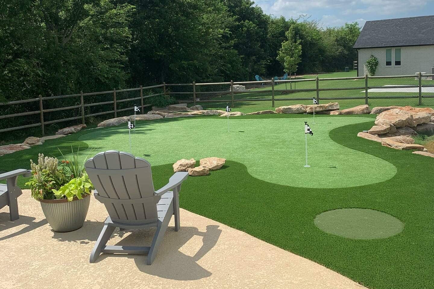 Backyard putting green with multiple flagsticks, border turf, rocks, fence, and patio chairs